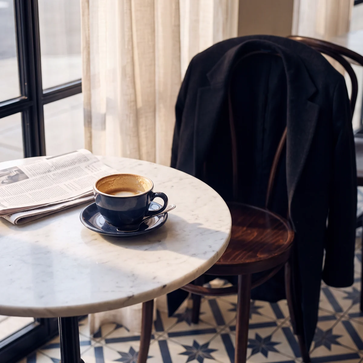 A round marble table by a curtained window with an open newspaper and a dark blue cup of coffee on its saucer, and a black coat over the back of a bentwood chair, on a floor of blue star-patterned tiles.