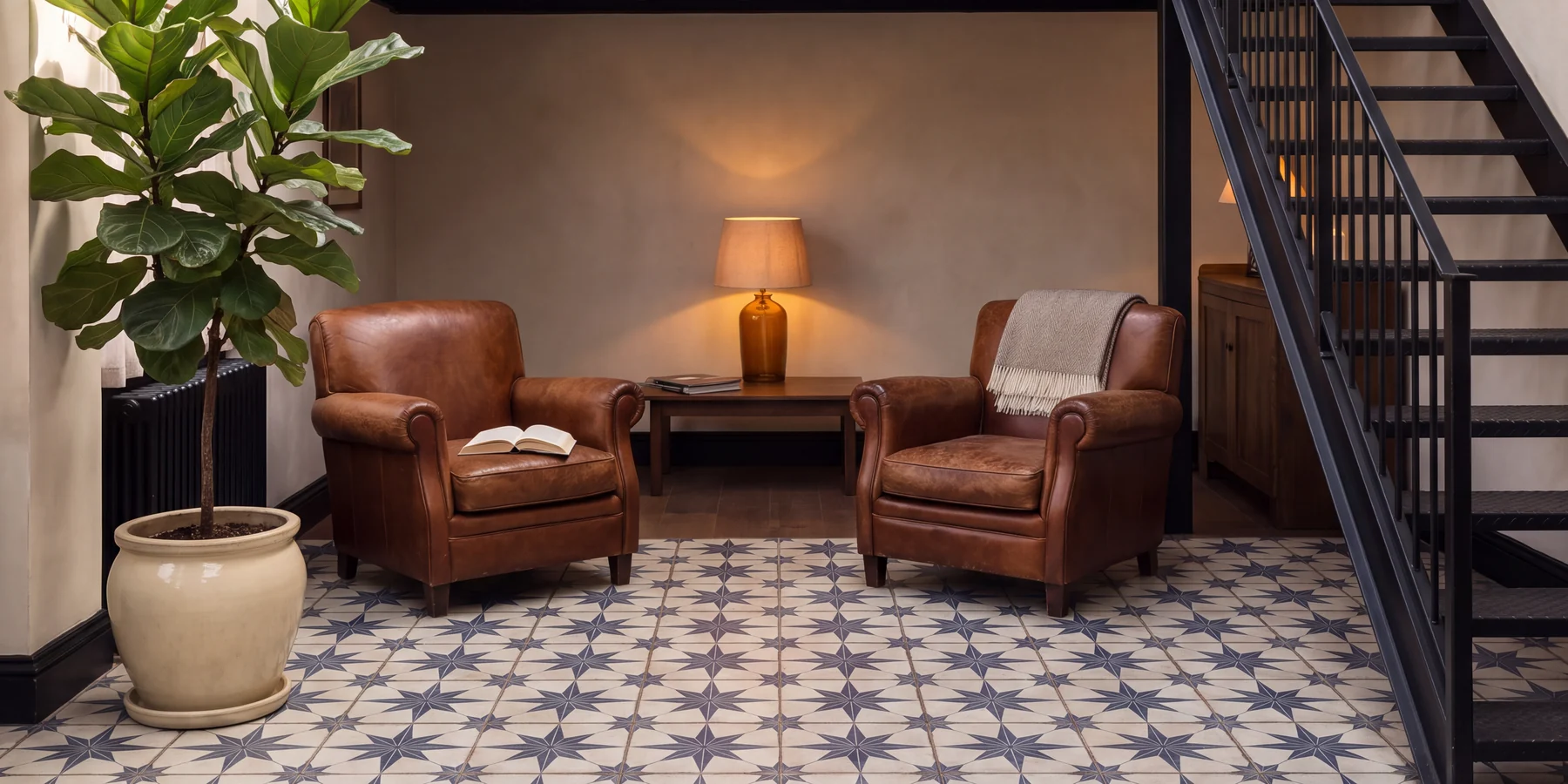 A reading corner: two brown leather armchairs facing a low wooden table with a lit amber lamp and folded newspapers on it, an open book on the left armchair and a cream throw over the right one, a fiddle leaf fig in a pale ceramic pot at the left edge, and a black steel staircase climbing off the right edge, on a floor of blue star-patterned tiles.